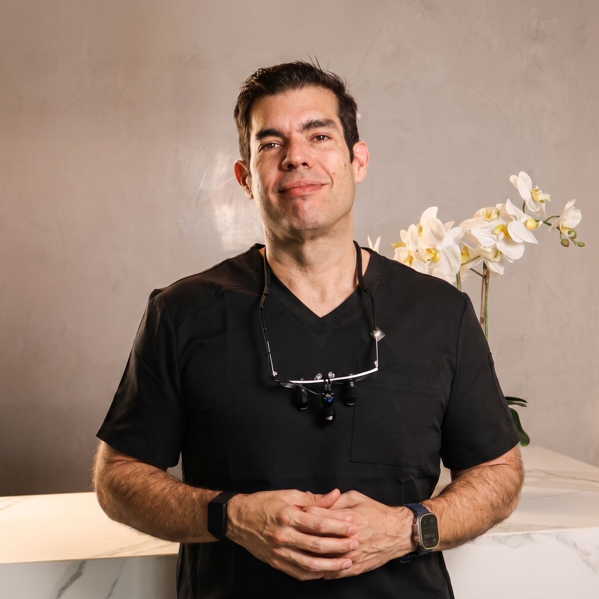 Dr. Carlos Aguilar León, implant and cosmetic dentist, standing in black scrubs with dental loupes at his clinic in Heredia, Costa Rica