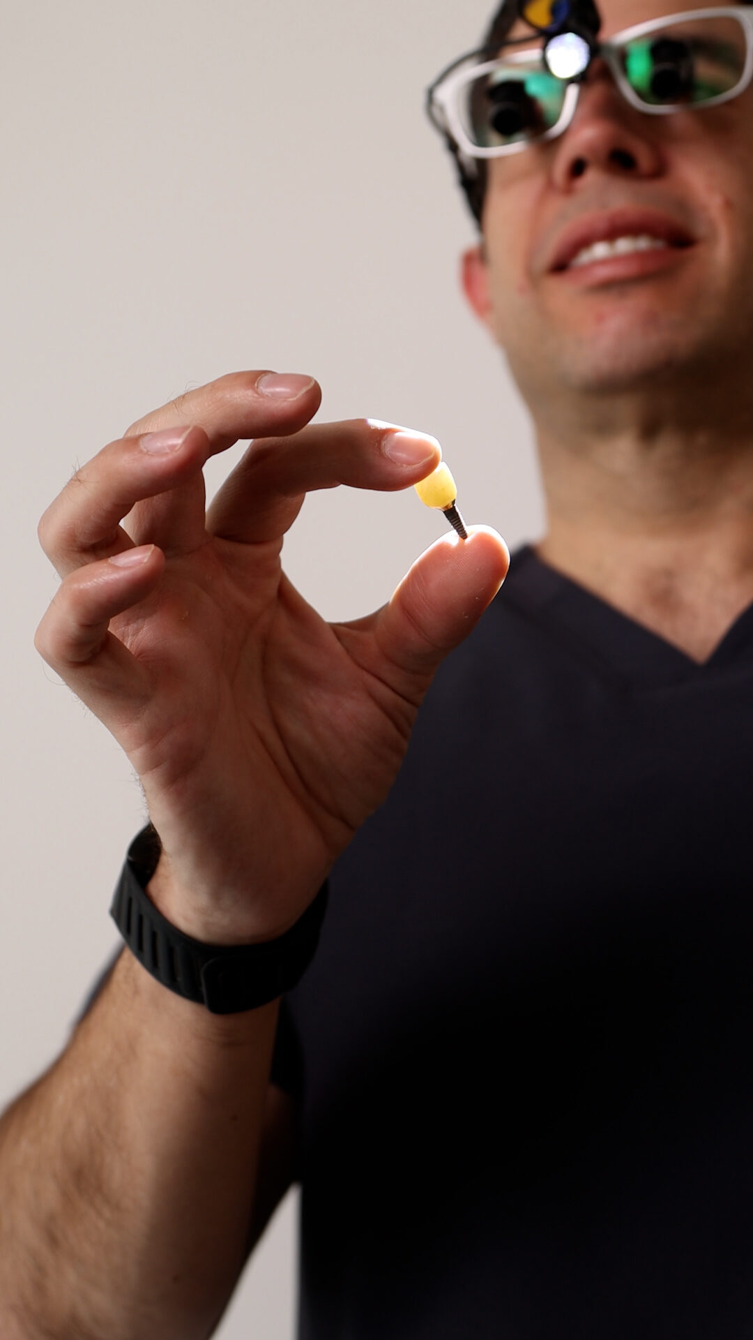 Dr. Aguilar, wearing surgical loupes, holding a single dental implant and its crown between his fingertips against the light